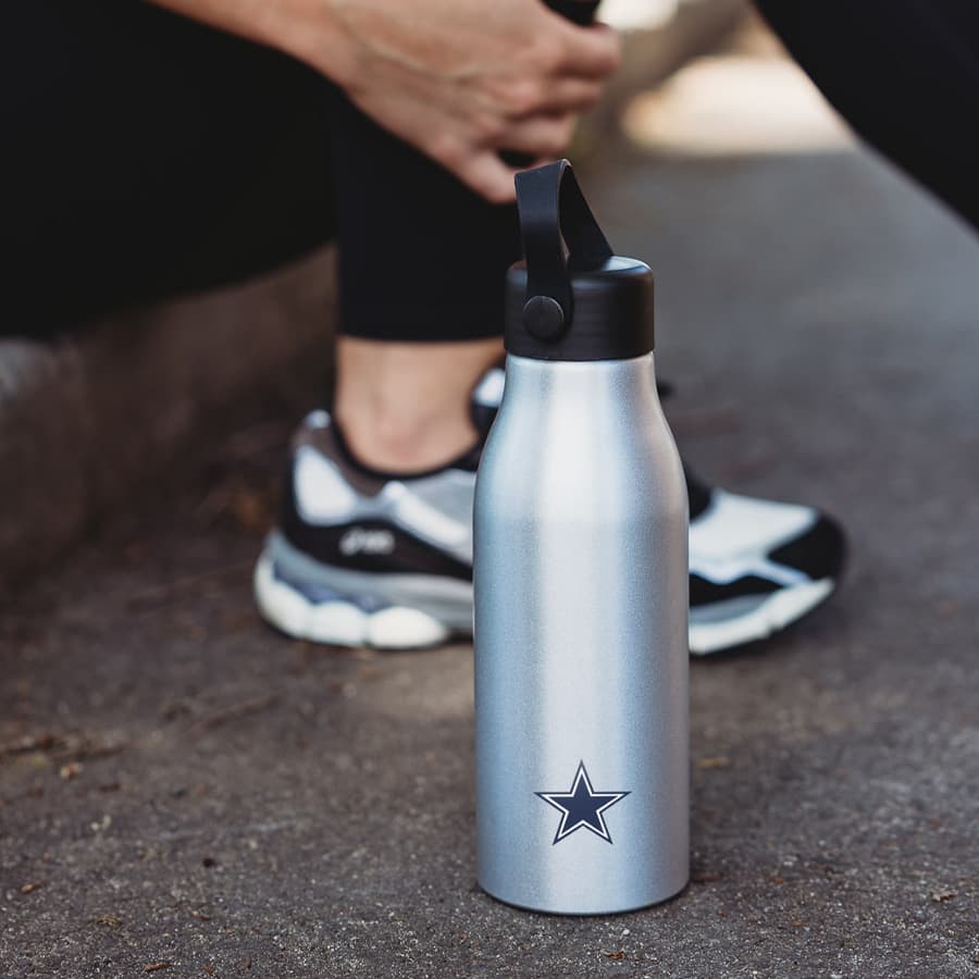 A drink bottle in silver with a logo, featuring a black lid and a sleek design, placed on a paved surface. Bentley Bottle 600ml - Image 1
