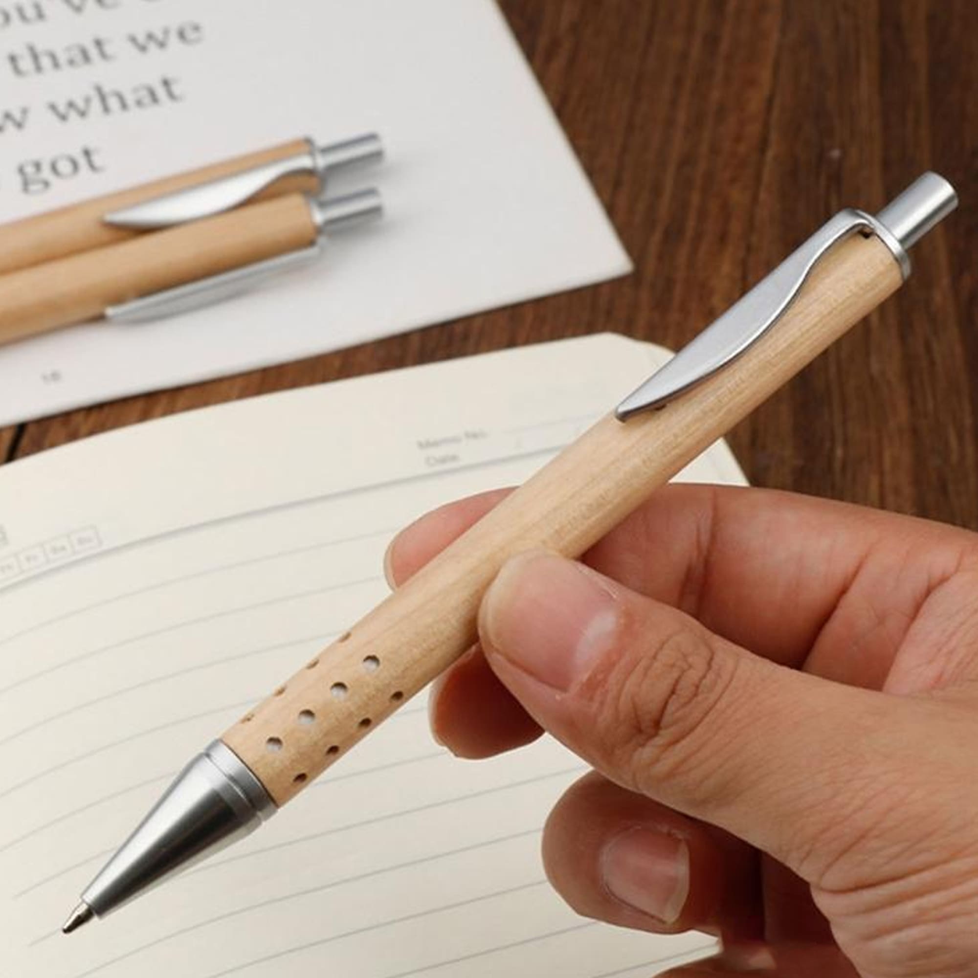 A pen with a wooden body and silver accents, held in a hand above a lined notebook. Pan Wood Pen - Image 1