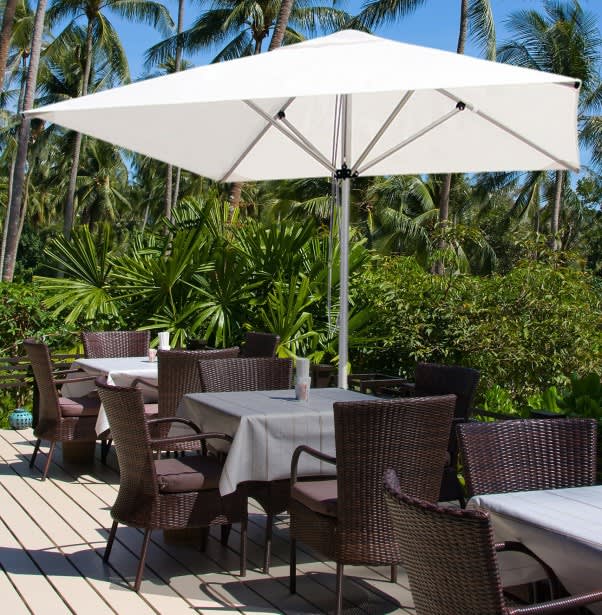 A white market umbrella without a valance positioned over outdoor tables and chairs surrounded by lush greenery. Market Umbrella No Valance 2m x 2m - Image 1