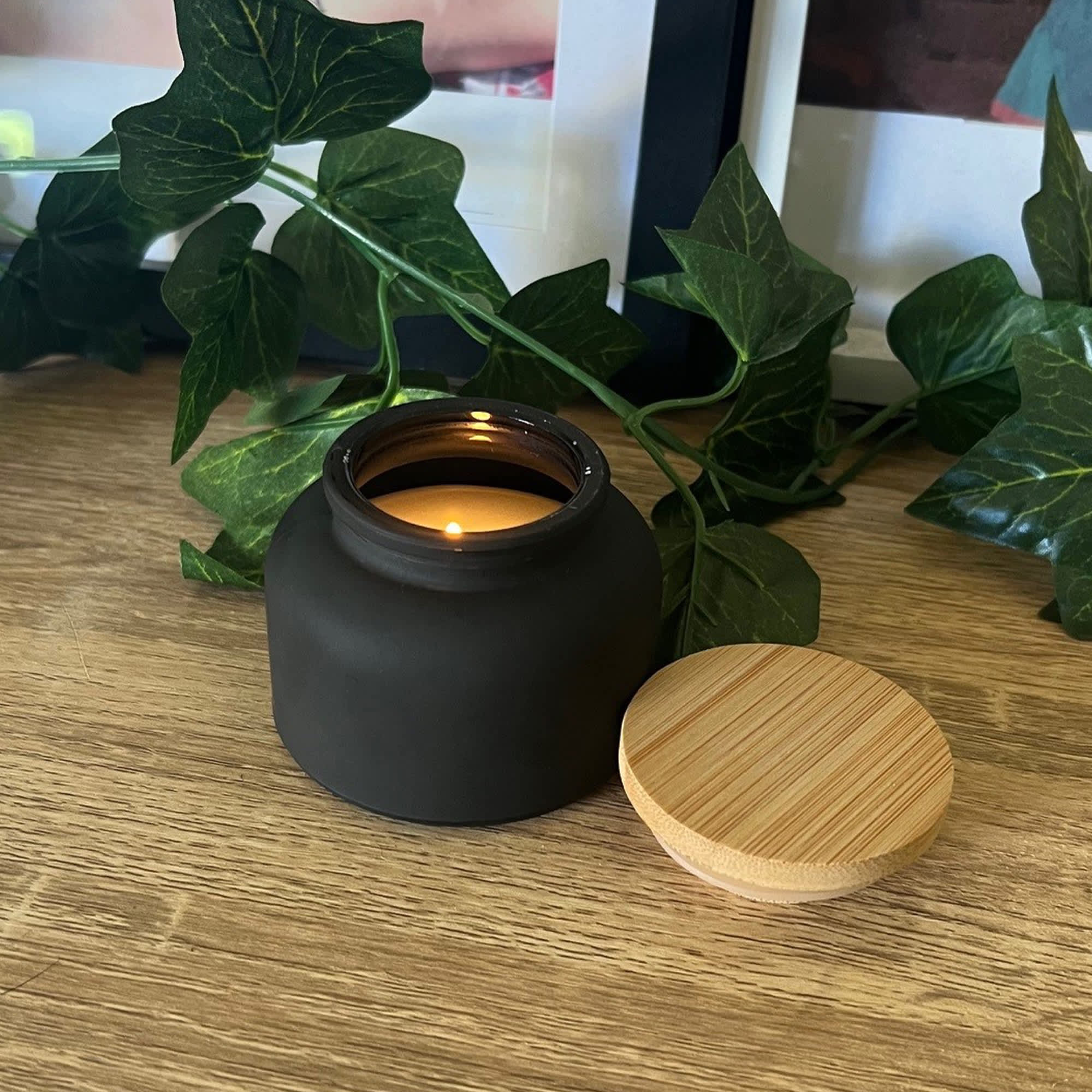 A black candle jar with a bamboo lid, placed on a wooden surface, surrounded by green ivy. Dream Candle - Image 1