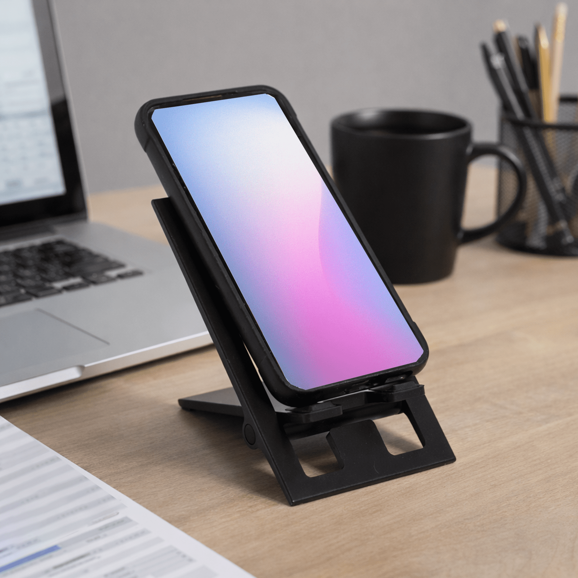 A black foldable phone stand with a phone displaying a gradient screen, set on a desk with a laptop and mug. The Felix Foldable Phone Stand - Image 1