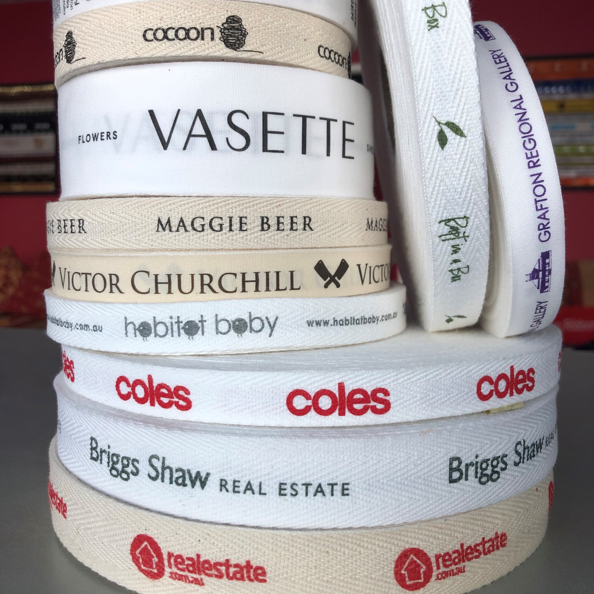 A stack of flat and herringbone cotton ribbons in cream and white, featuring various logos and text designs. Custom Printed Flat and Herringbone Cotton Ribbon 10mm - Image 1
