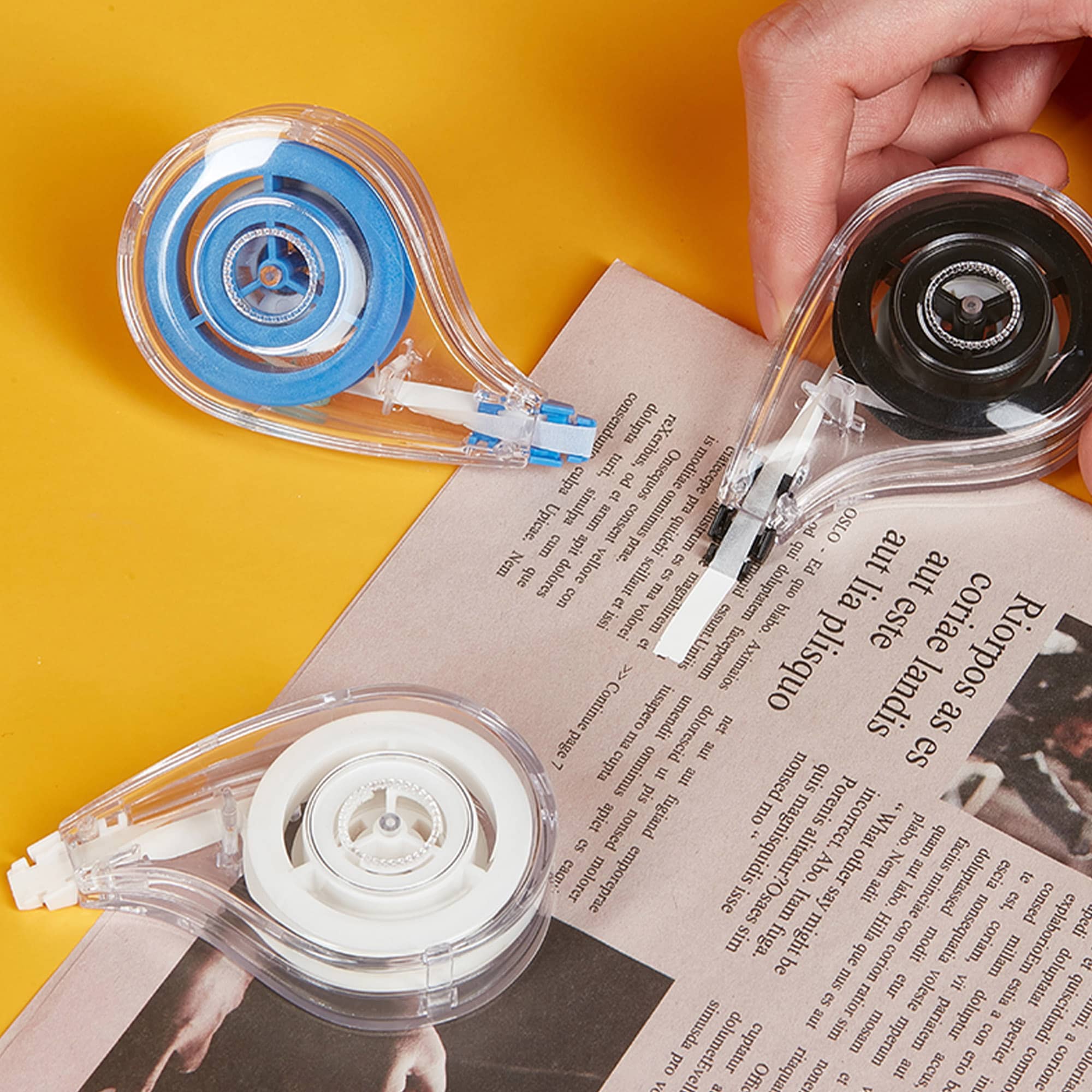 Three correction tape dispensers in blue, white, and black, placed on a magazine against a yellow background. Wigo 8m Correction Tape - Image 1