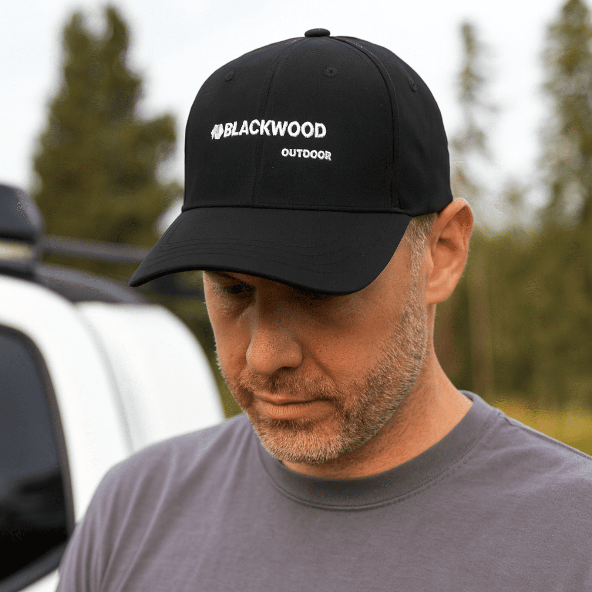 A black strapback baseball cap with a logo, worn by a person outdoors near a vehicle. Poly Strapback Baseball Cap - Image 1