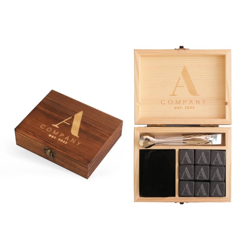 A wooden box featuring a logo, containing nine black whiskey stones and two stirring utensils. Whiskey Stones Set - Image 1