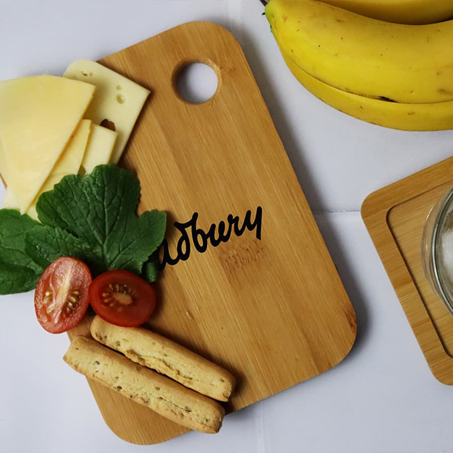A small bamboo cutting board with cheese, tomatoes, mint leaves, and snacks, featuring a logo, beside ripe bananas. Bamboo Cutting Board (Small) - Image 1