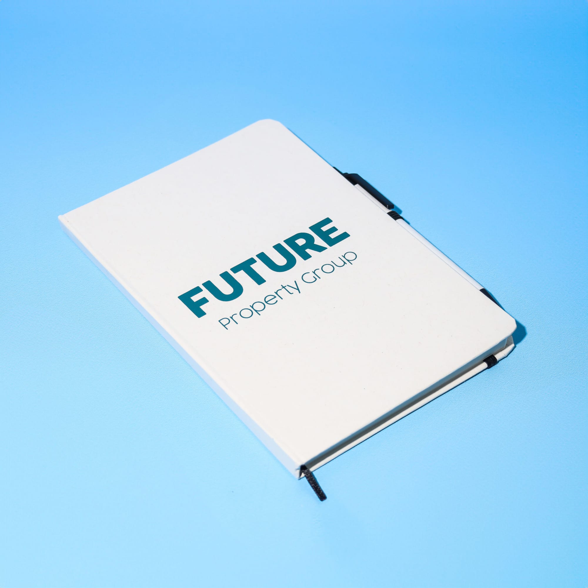 A white A5 notebook with a logo and a black pen resting on a blue background. Milky A5 Notebook & Pen Set - Image 1
