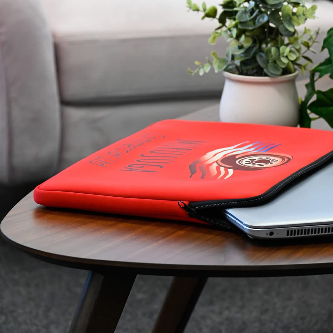 A red laptop sleeve featuring a logo, resting on a wooden table beside a plant and a laptop. Bowles Laptop Sleeve - Image 1