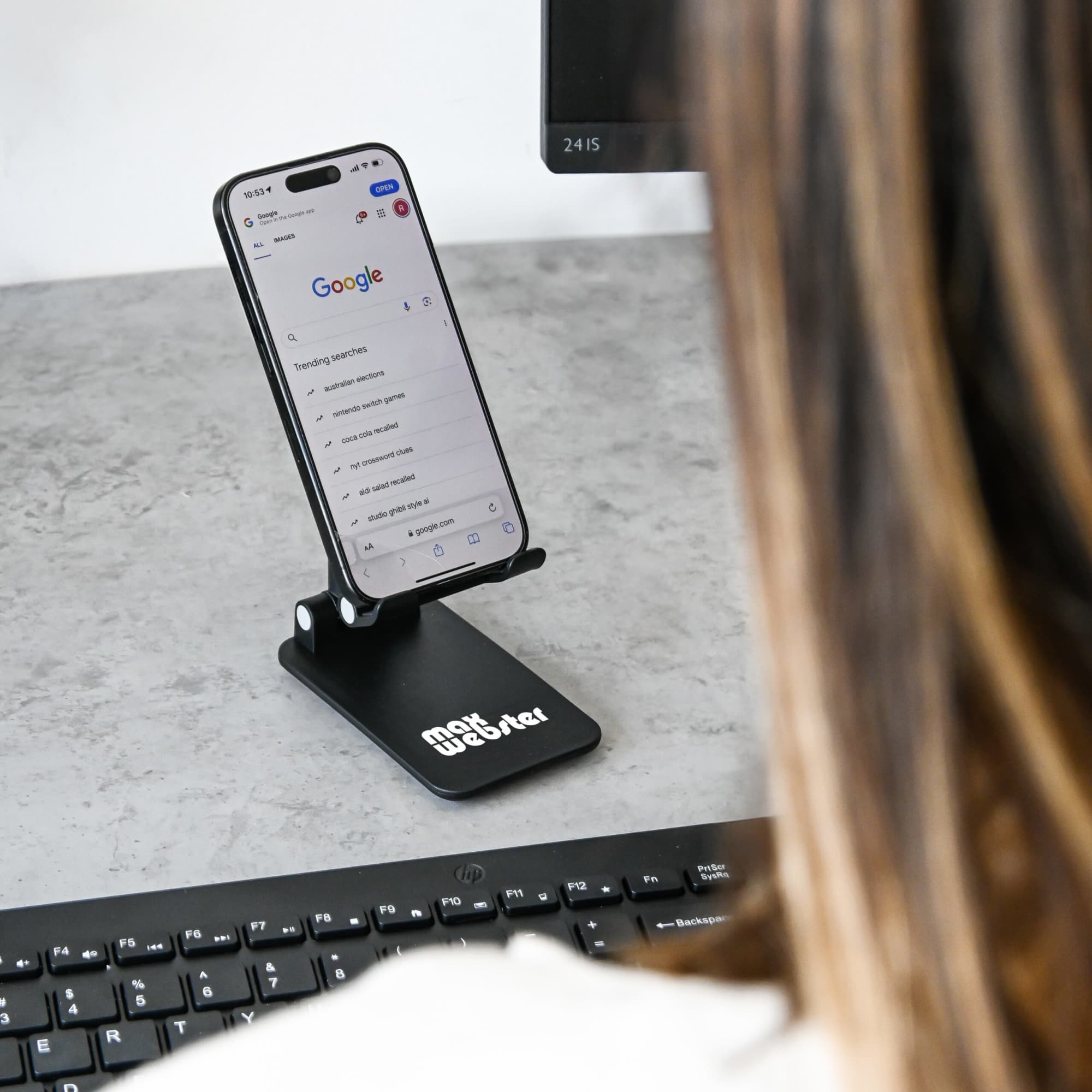 A black folding desktop mobile phone stand holding a smartphone, positioned on a desk near a computer keyboard. Folding Desktop Mobile Phone Tablet Stand - Image 1
