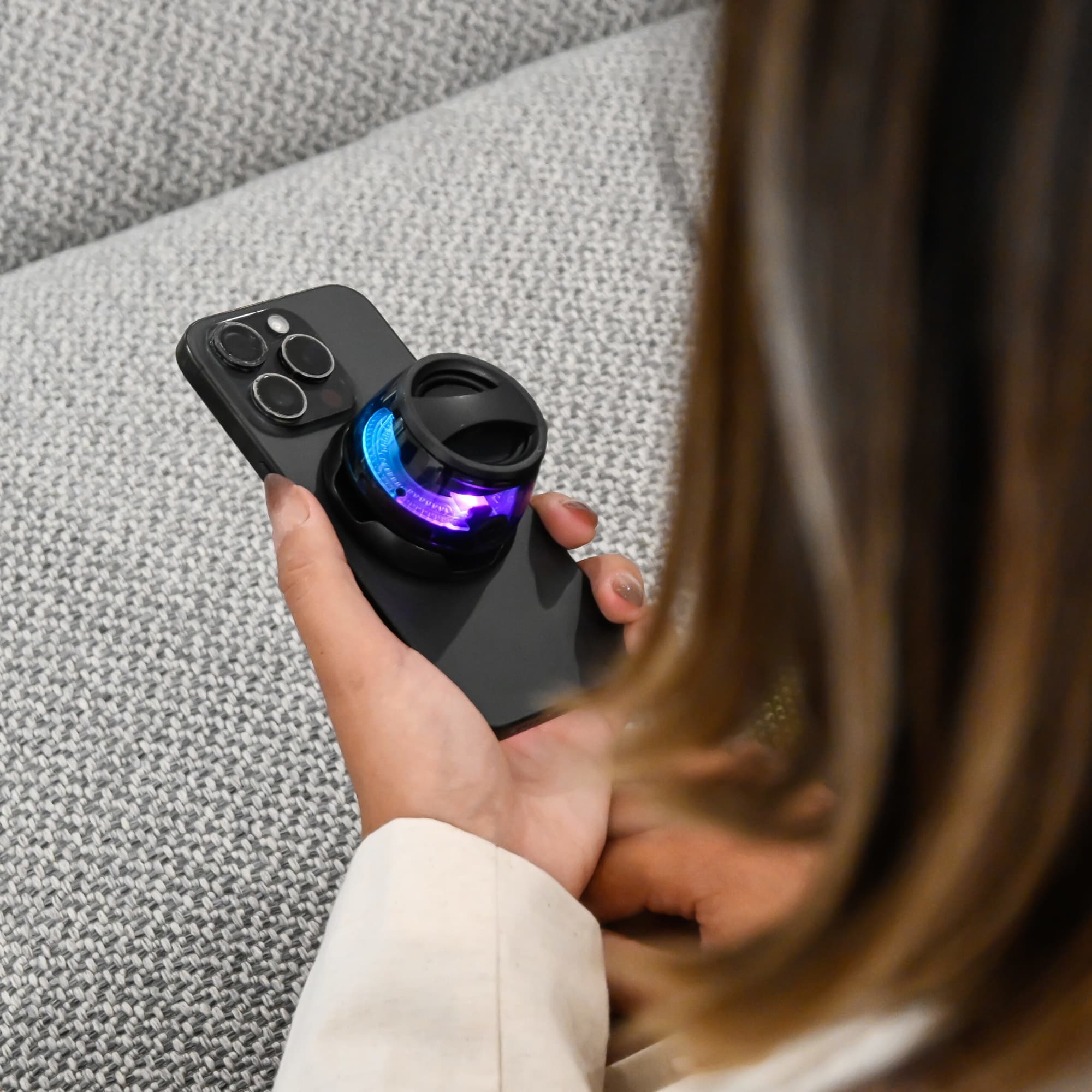 A compact, black speaker with colourful lights rests on a smartphone in a person's hand. The background features a textured fabric. Bentley Brook Speaker - Image 1
