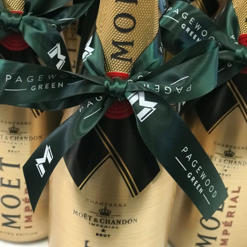 A gold champagne bottle wrapped with a dark green custom printed satin ribbon featuring a logo and text. Custom Printed Double Sided Woven Edged Satin Ribbon 24mm - Image 1