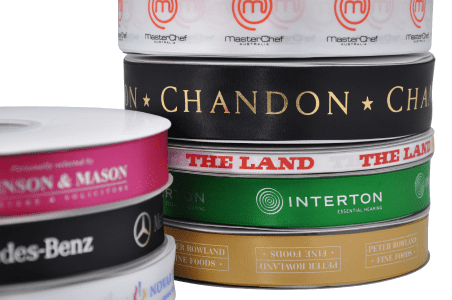 A stack of various custom printed double-sided satin ribbons in colours like black, pink, white, and green, with logos. Custom Printed Double Sided Woven Edged Satin Ribbon 7mm - Image 1