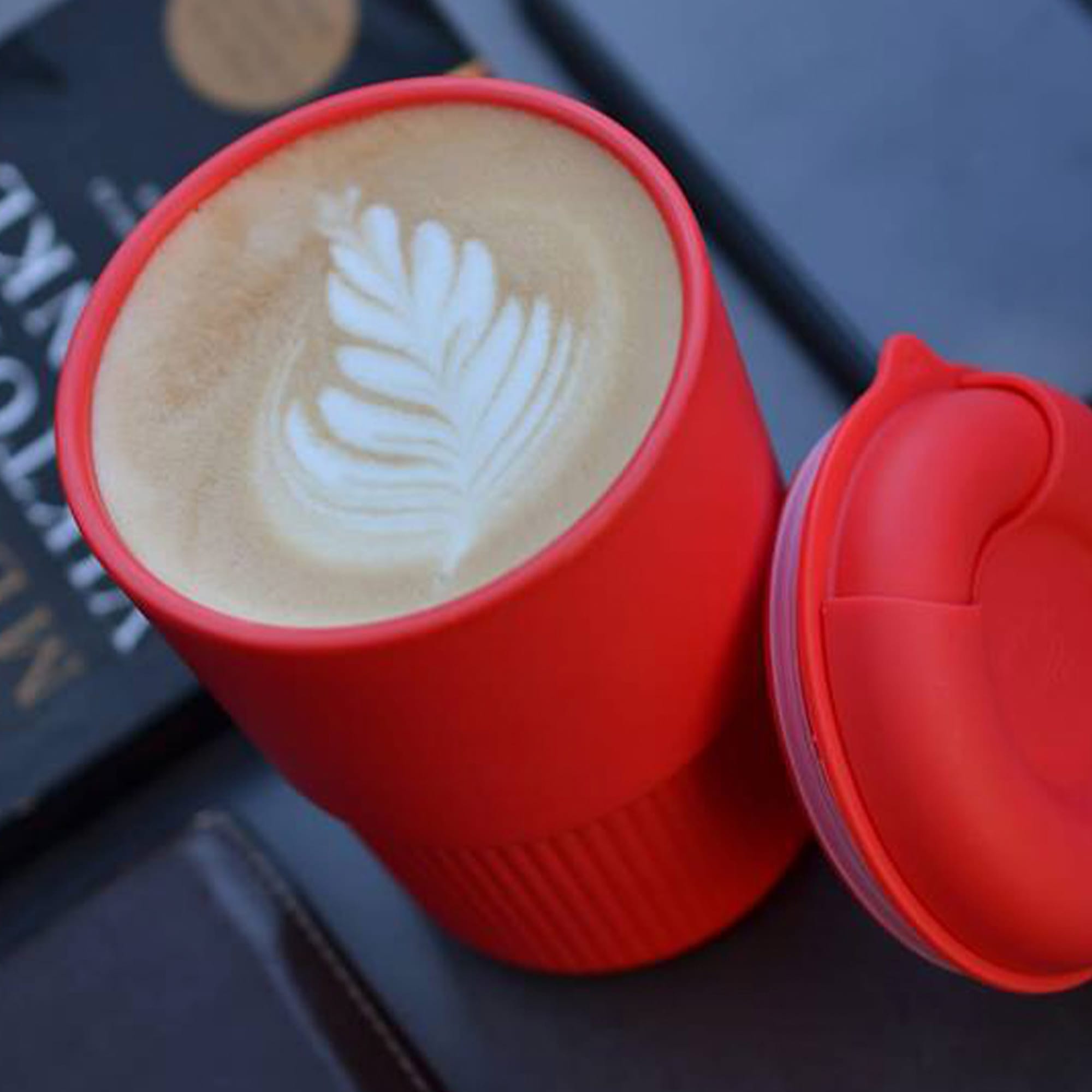 A reusable coffee cup in bright red with a latte featuring a leaf pattern, accompanied by a matching lid. Ecco Kuppa Universal - Image 1