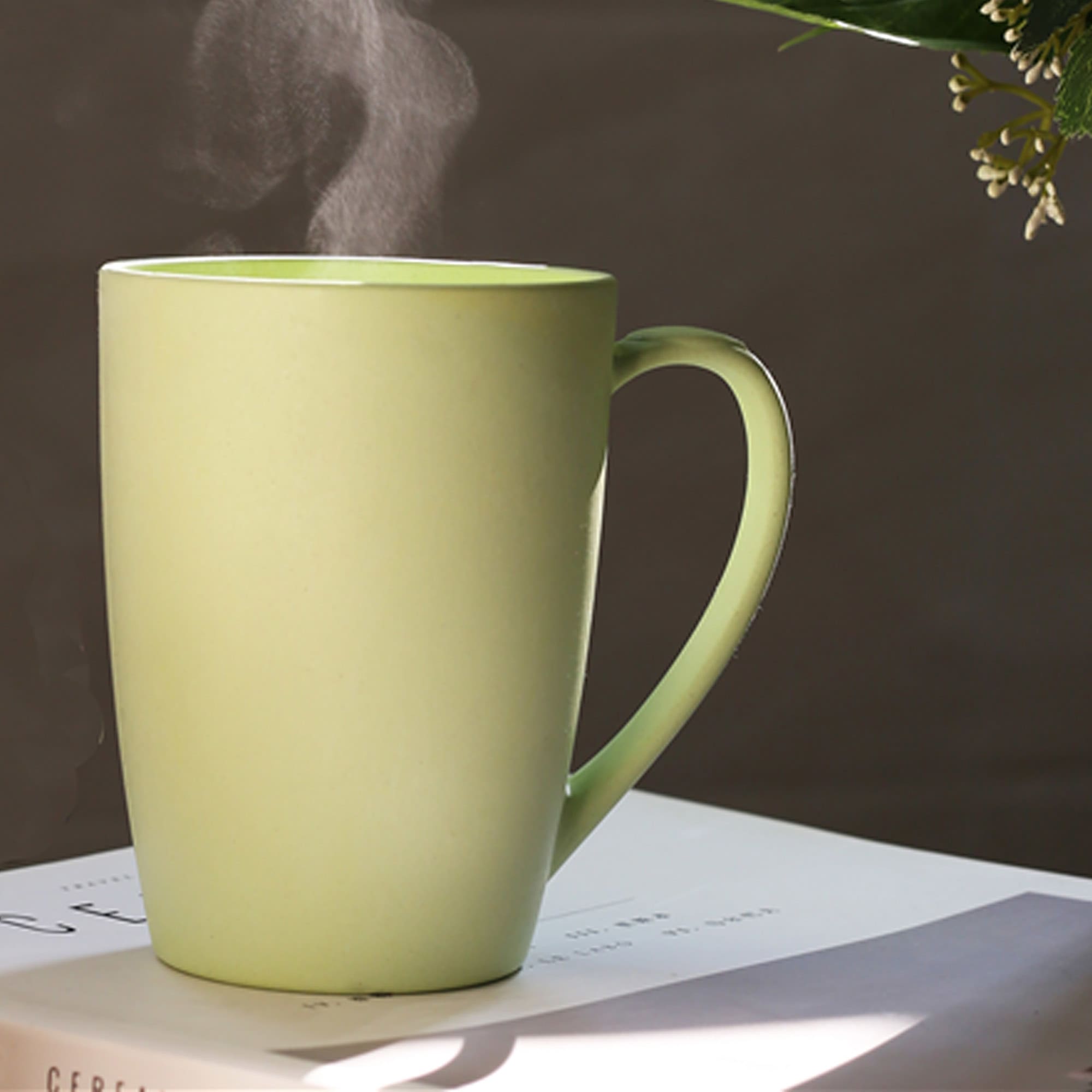 A reusable bamboo mug in a soft green colour, emitting steam, sits on a book with foliage in the background. Envee Bamboo Mug - Image 1