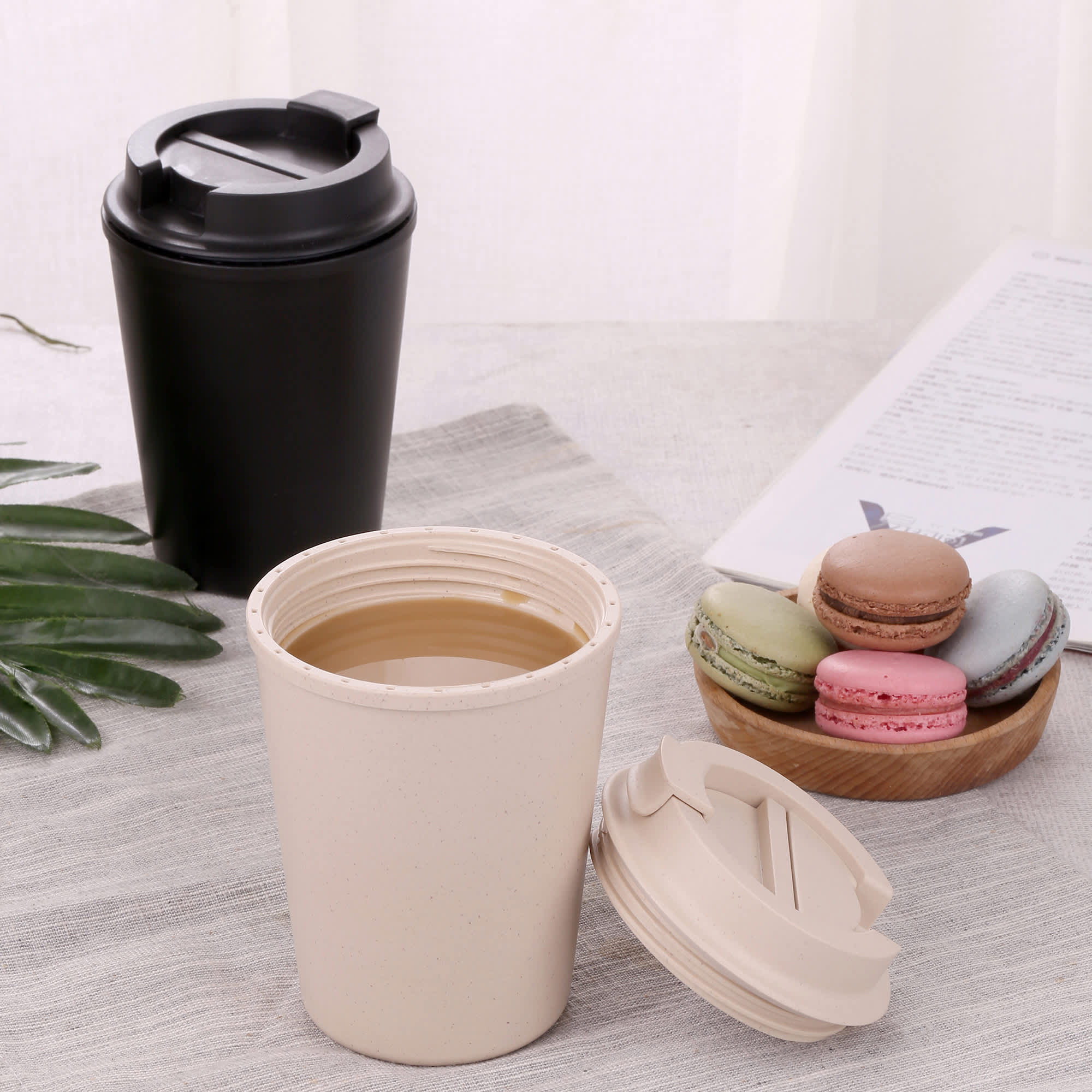 A reusable coffee cup in beige with a lid, next to a black cup and a plate of colourful macarons. Karvo Ecco Kuppa - Image 1