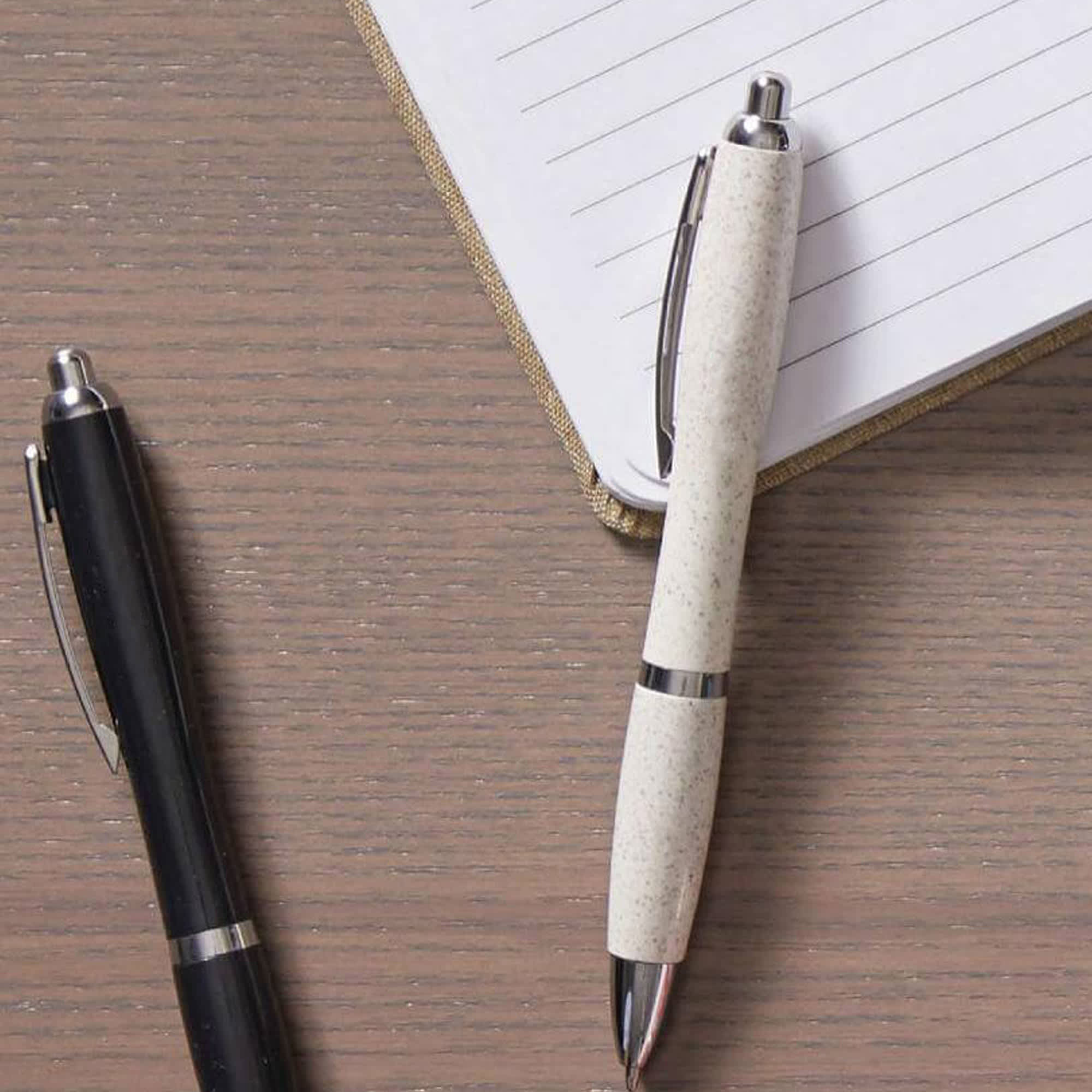 Two plastic pens rest on a wooden surface beside a notepad. One pen is black, and the other is white with a speckled design. Blast Eco Pen - Image 1