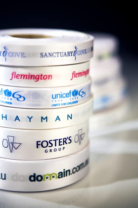 A stack of white ribbons featuring various logos and text, prominently displaying custom full colour printing. Custom Full/Exact Colour Print on White Ribbon 36mm - Image 1