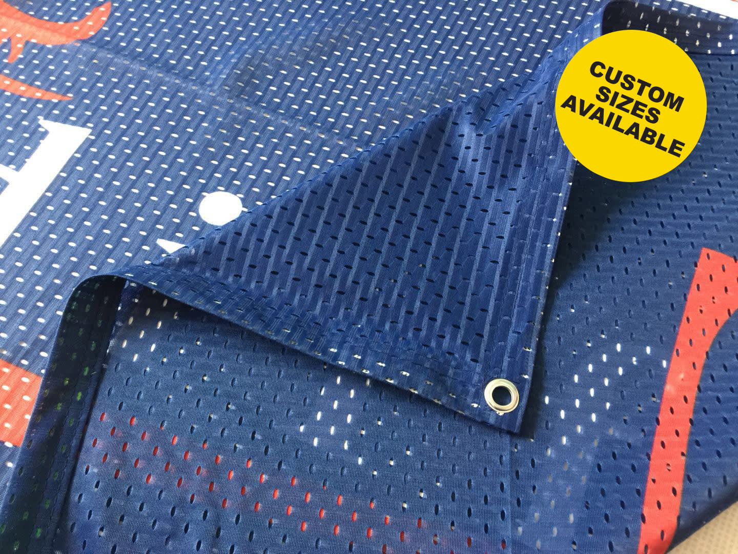A mesh polyester banner with a blue background and perforated design, featuring a corner grommet and branding. Mesh Polyester Banner - Image 1