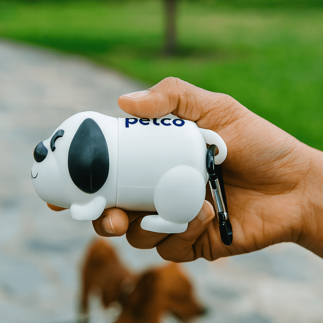 A white dog-shaped waste bag dispenser with black details, held in a hand, featuring a logo. Dog Waste Bag Dispenser - Image 1