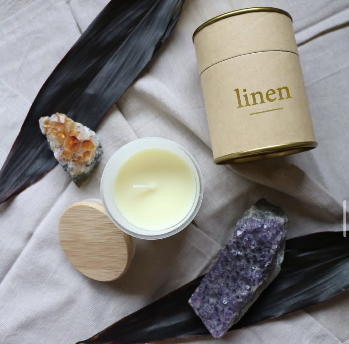 A large Relax Candle in a gold and kraft cylindrical container, surrounded by crystals and dark leaves on a fabric surface. Relax Candle - Large - Image 1