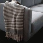 A light grey and white striped throw blanket with tassels draped over the arm of a blue sofa.