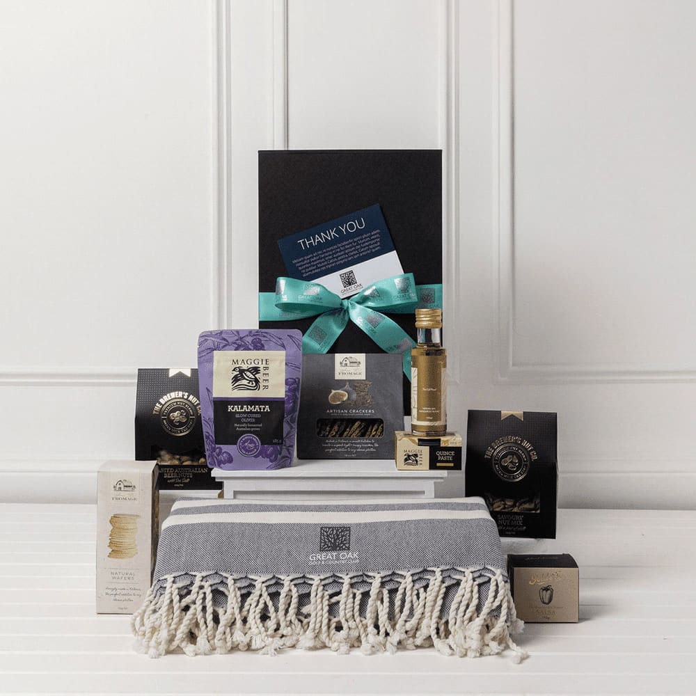 A gourmet gift pack featuring assorted food items, a stylish grey throw, and a bottle, all presented in a black box. The Gourmet Retreat Gift Pack - Image 1