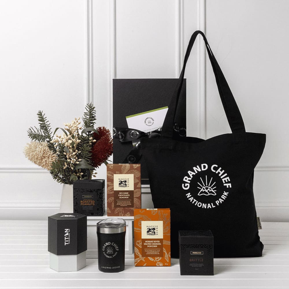 A black tote bag with a logo, surrounded by various sweet treat boxes, a candle, and decorative dried flowers. Custom Sweet Treats & Essentials Gift Pack - Image 1