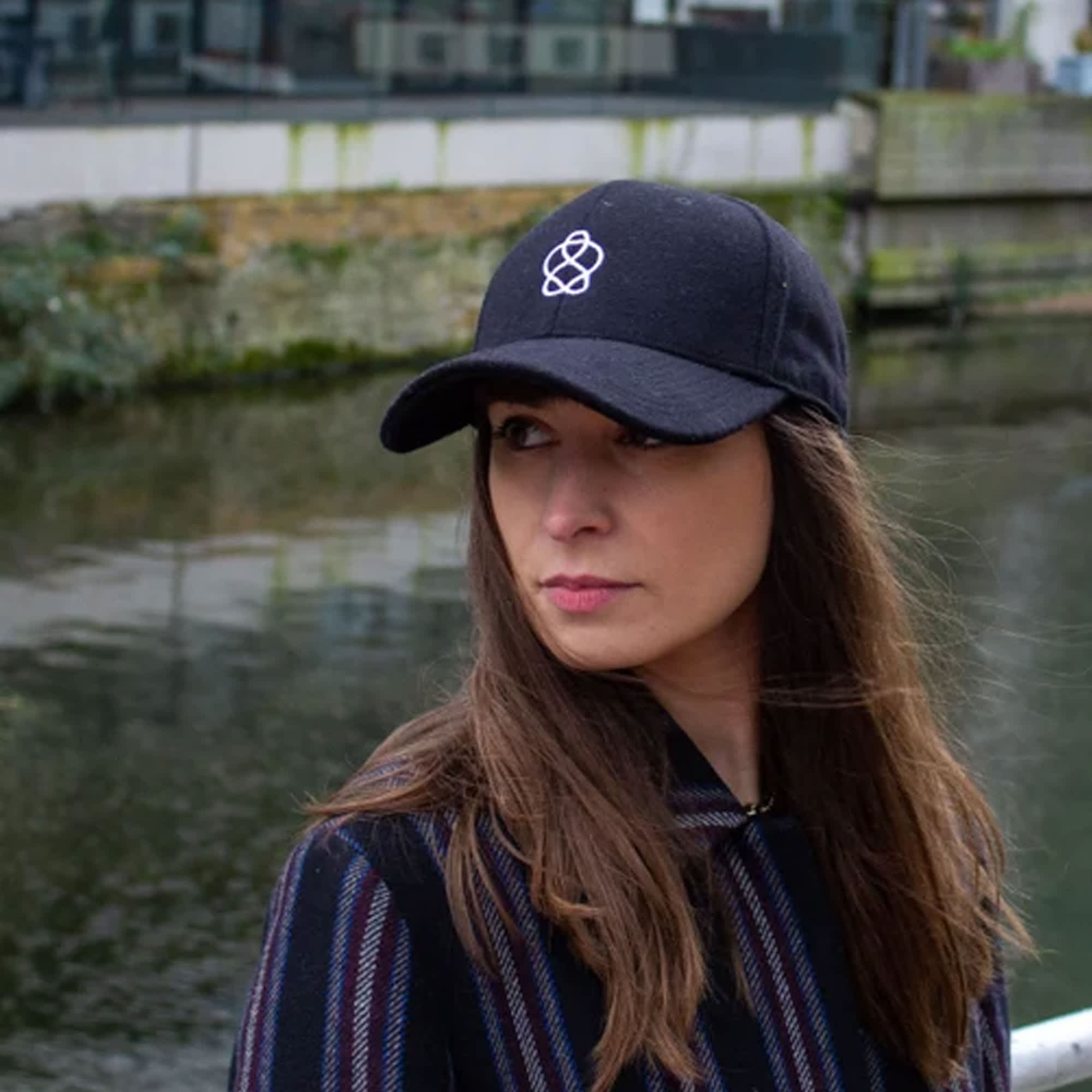 A black heavy acrylic cap featuring a logo, worn by a person beside a body of water. Orega 6 Panel Heavy Acrylic Cap - Image 1