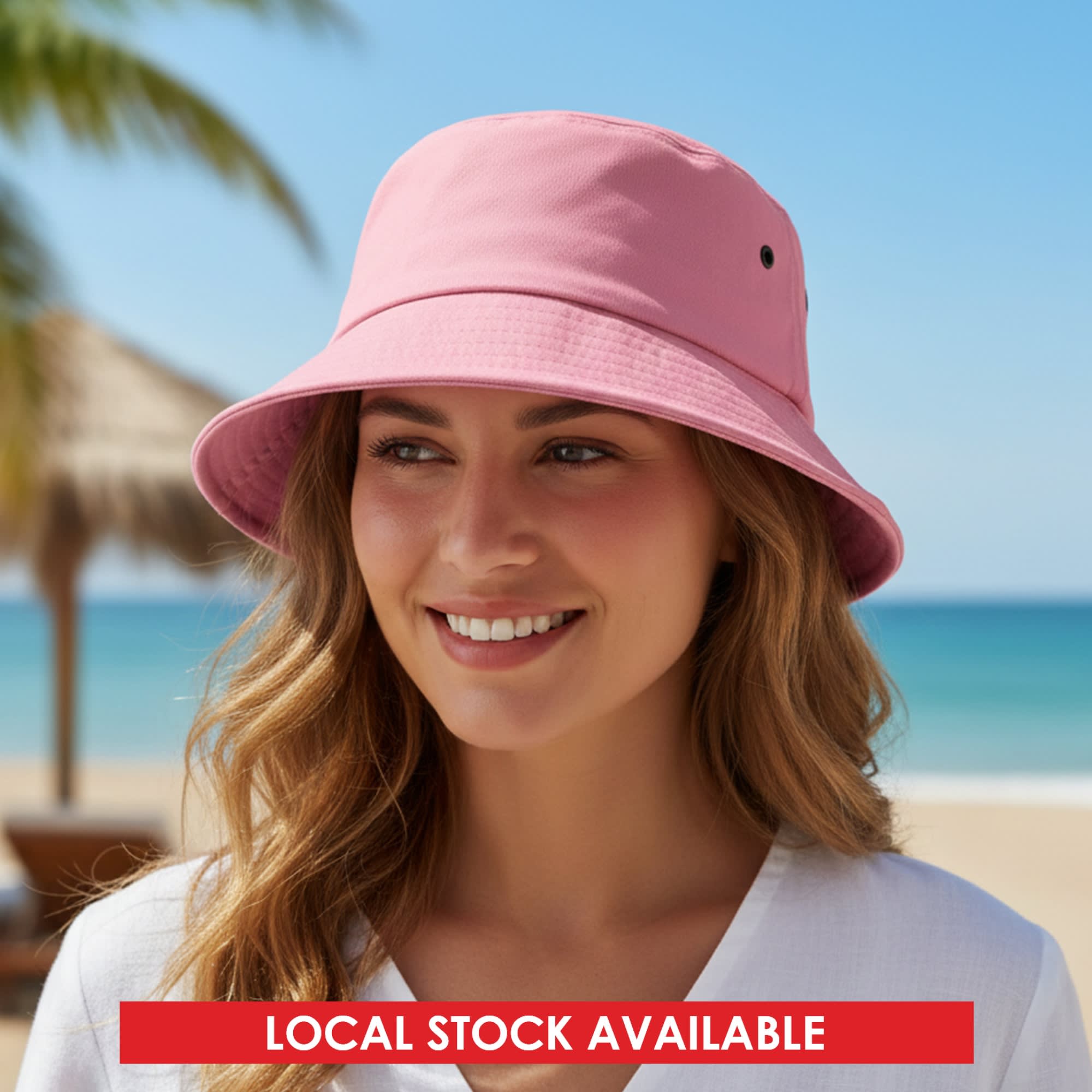 A pink bucket hat sits on a smiling woman's head with a beach backdrop and palm trees. The hat features a logo. Long Beach Bucket Hat – Medium - Image 1