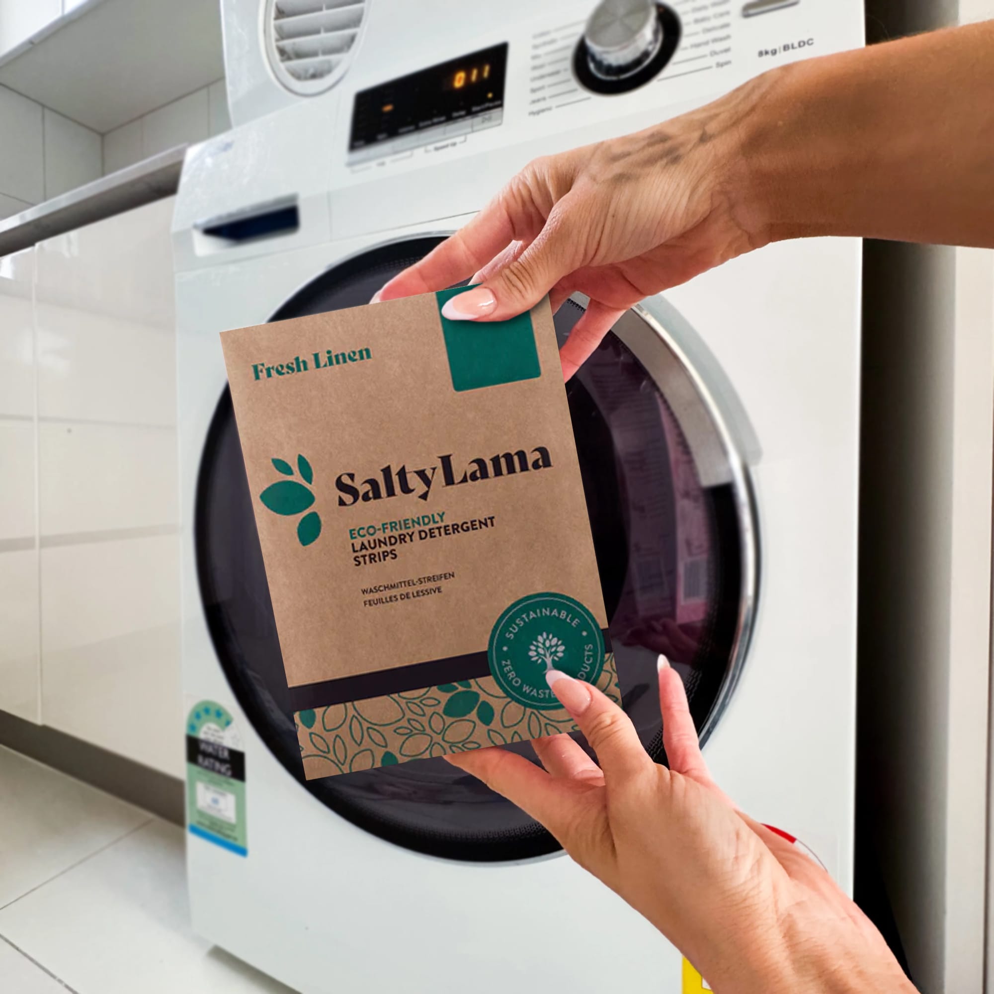 Eco-friendly laundry detergent strips in brown packaging with a logo, held in hands near a washing machine. Laundry Detergent Sheets - Image 1