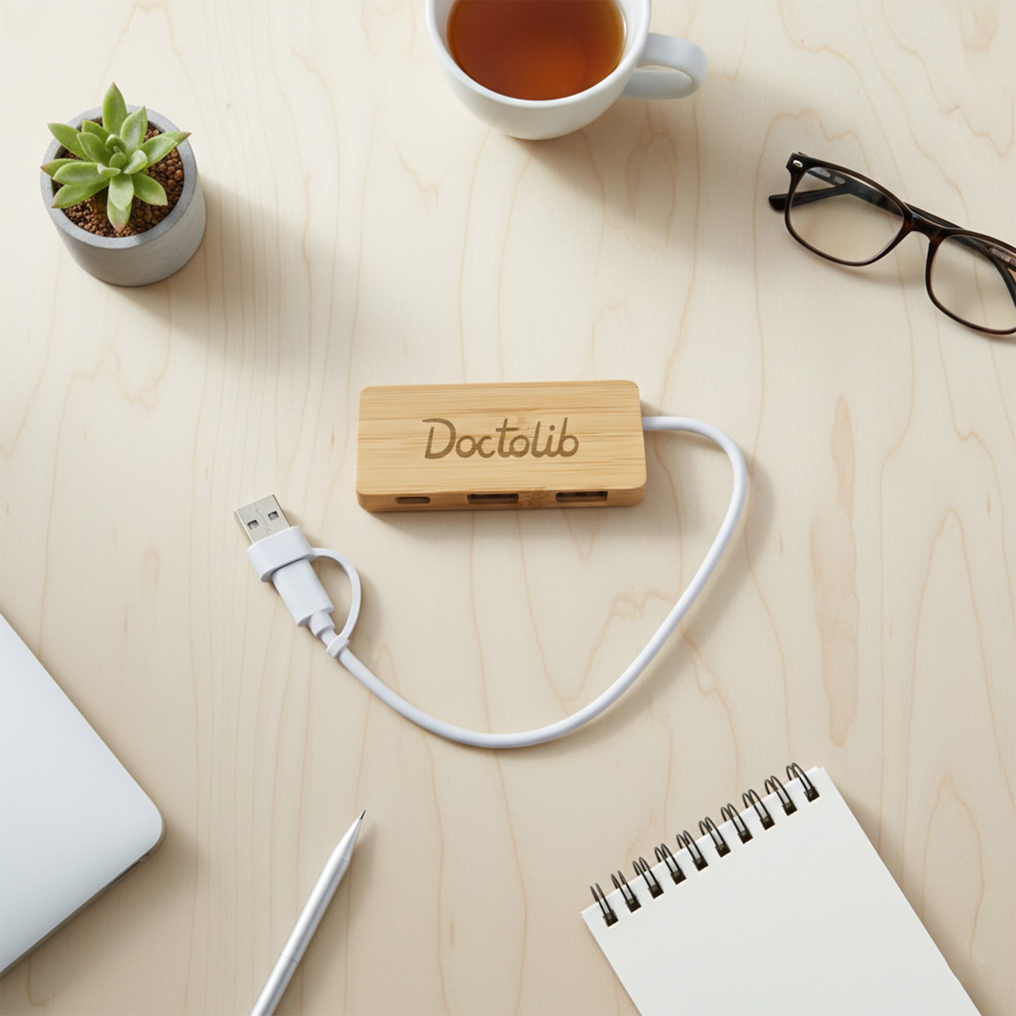 A bamboo USB hub with a white cable, placed on a wooden surface alongside a cup, notebook, and glasses. Bamboo USB Hub - Image 1
