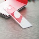 A transparent red 15cm magnifying ruler lying on a wooden surface beside a notebook. It has a brand logo.