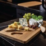 A bamboo cheese board features a variety of cheeses, grapes, nuts, and a cheese knife, with a sleek black countertop.