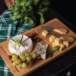 A bamboo cheese board with a circular cheese, assorted cheeses, grapes, and two cheese knives with wooden handles.