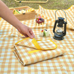 A folded yellow and white checkered picnic blanket lies on grass, accompanied by a lantern and picnic items.