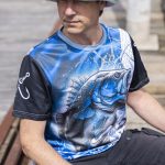A fishing tee featuring a blue and black design with a graphic of a fish, worn by a person outdoors.