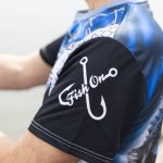 A men's fishing tee in blue and black with a fishing hook design on the sleeve and logo branding.