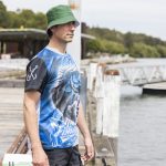 A man wearing a blue and black polyester sublimated fishing tee and a green bucket hat by the water.