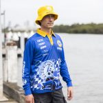 A man wearing a blue long sleeve fishing polo with a yellow collar and a matching bucket hat, beside a waterway.
