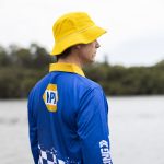 A man wearing a blue long sleeve fishing polo and a yellow bucket hat stands by the water. The shirt features a logo.