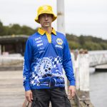 A man wearing a blue long sleeve polyester fishing polo with a graphic design and a yellow bucket hat.