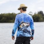 A man wearing a blue long sleeve polyester fishing tee with a fish graphic, standing by a body of water.