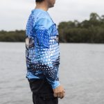 A long sleeve sublimated fishing tee in blue, featuring a fish design, displayed against a waterfront backdrop.