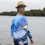 A man wearing a long sleeve blue polyester fishing tee, featuring a fish graphic, near a body of water.