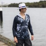 A man stands by the water wearing a navy blue and grey patterned 1/4 zip fishing hoodie and a white hat.