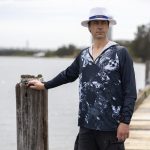 A man stands by the water wearing a navy and white polyester sublimated 1/4 zip fishing hoodie, with a hat and shorts.