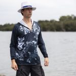A man in a navy and white patterned polyester fishing hoodie stands by the water, wearing a white hat.