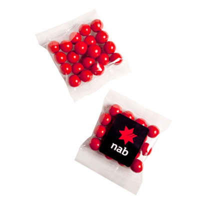 A clear packet containing red chocolate-coated treats and a small black square with a logo. Orange Choc Drops (Jaffa look alike) 50g - Image 1
