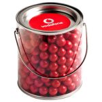 A clear bucket filled with vibrant red chocolate balls, featuring a lid and a metal handle.