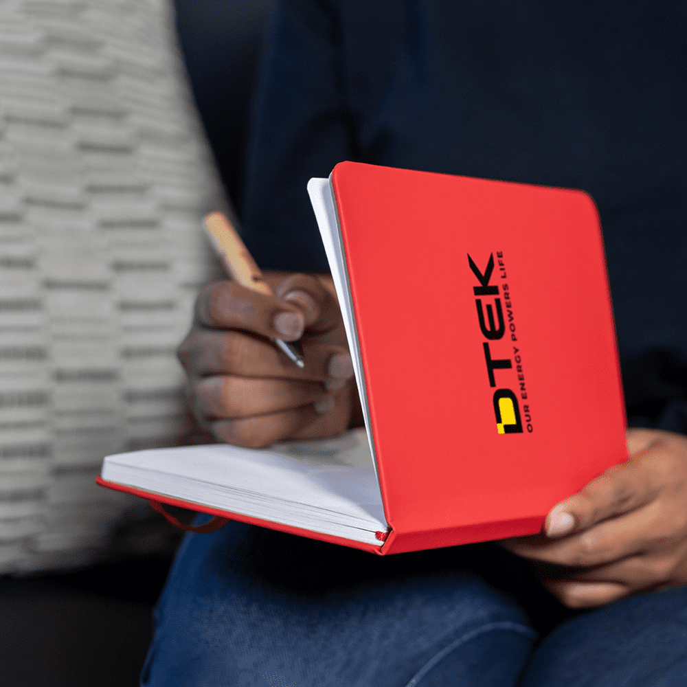 A red A5 notebook with a logo on the cover, held open with a hand writing inside. Space A5 Notebook - Image 1